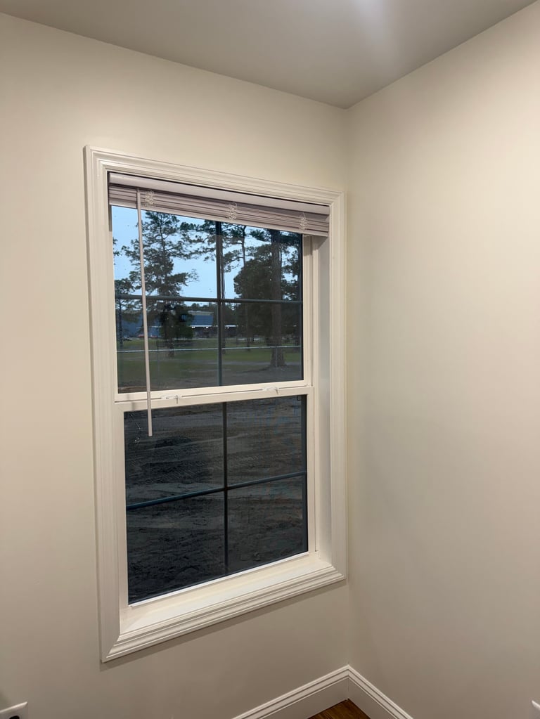 Double-hung window with white frame on beige wall, showing view of trees, lawn, and outdoor scenery