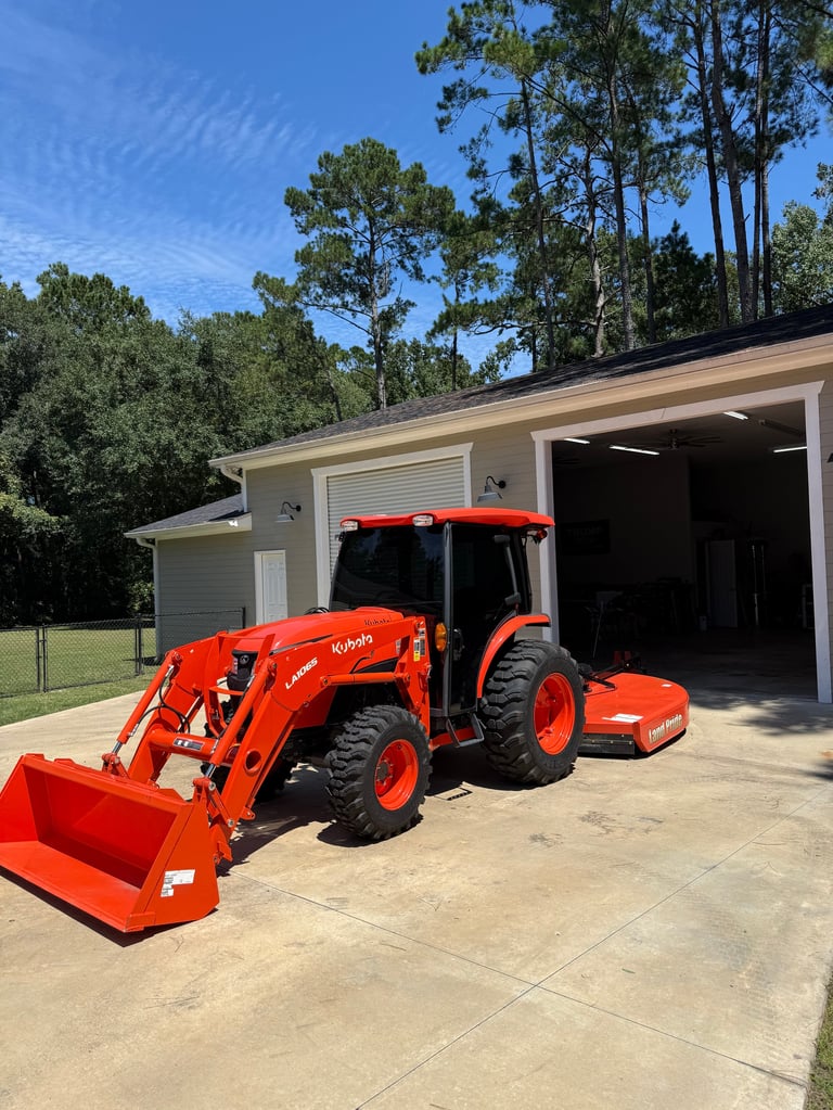 Red compact tractor with front loader bucket parked in driveway beside gray storage building and pine trees